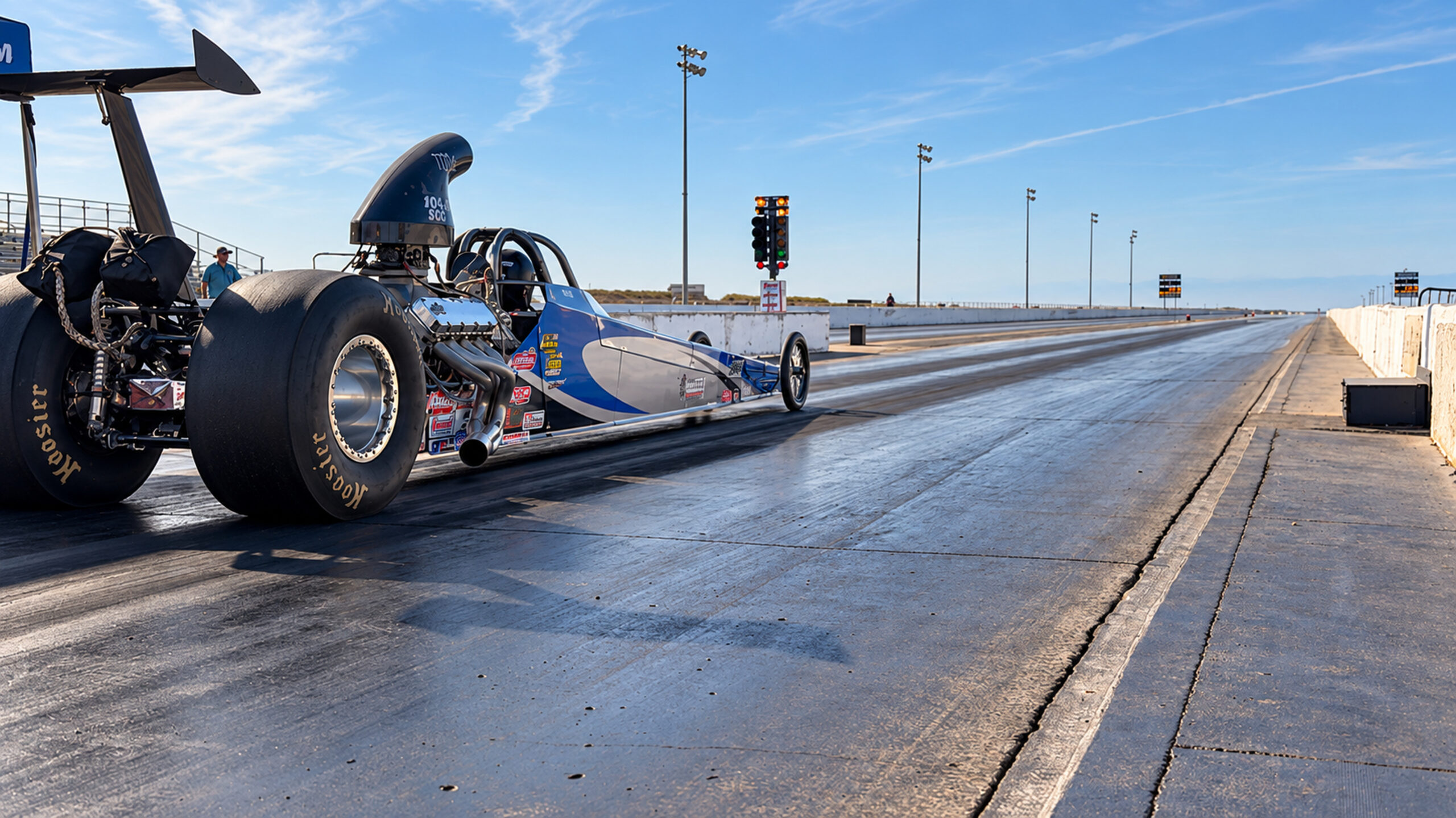 Rear view of a blue and silver Super Comp dragster at the starting line, with the Christmas Tree and drag strip ahead.